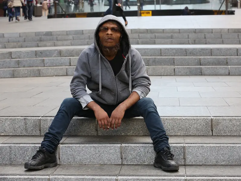 actor in prosthetic makeup of ancient human sits on public steps wearing a hoodie